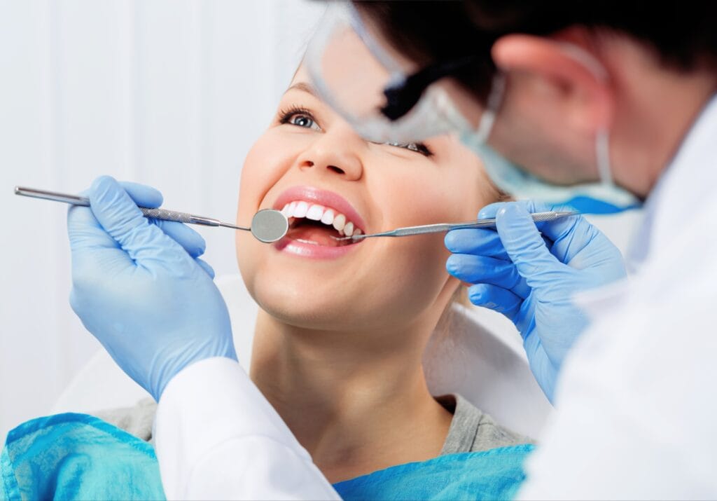 Smiling woman receiving a dental cleaning from a dentist using dental instruments, including a mirror and scaler.