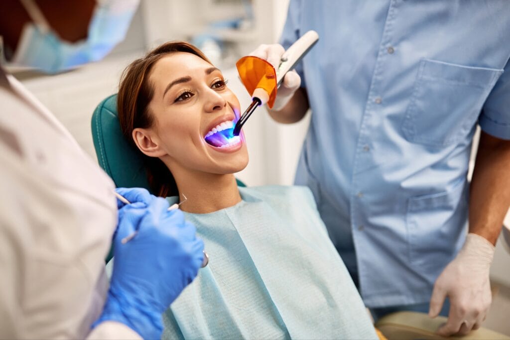 Woman receiving a dental filling procedure in a clinic, with a curing light being applied to her teeth by a dentist wearing gloves and protective equipment.