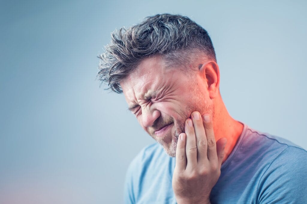 Middle-aged man with graying hair holding his cheek in pain and wincing, showing signs of a severe toothache or dental emergency against a neutral background.