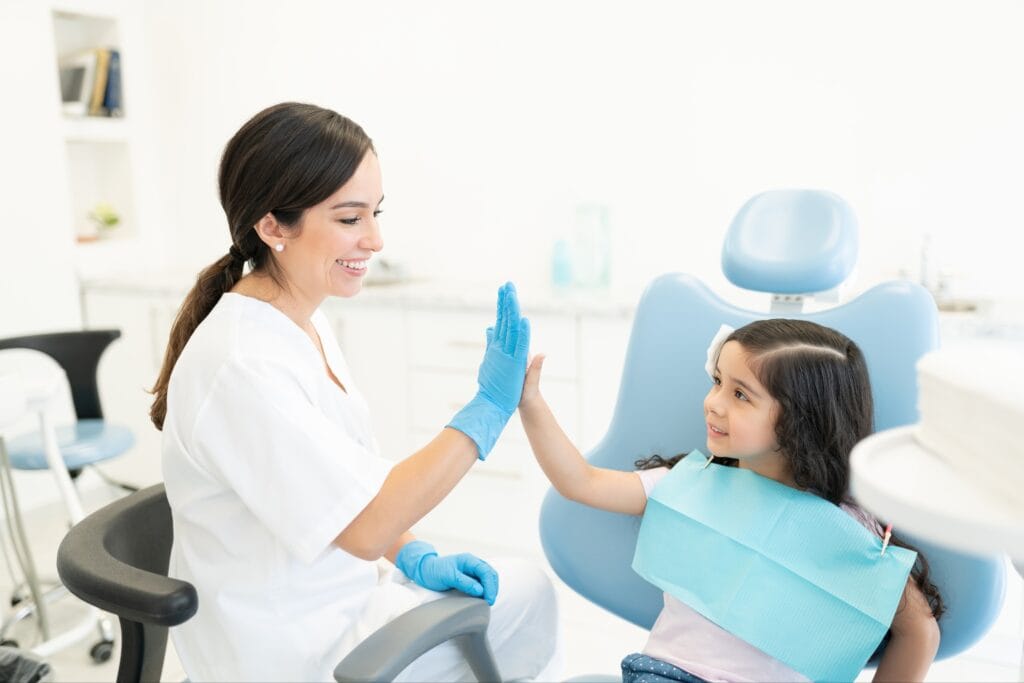 Smiling female dentist giving a high-five to a young girl sitting in a dental chair, celebrating a successful first dental visit in a bright, modern clinic.