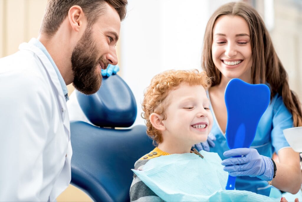 Smiling young boy sitting in a dental chair looking at his teeth in a blue tooth-shaped mirror, with a male dentist and female dental assistant happily observing.