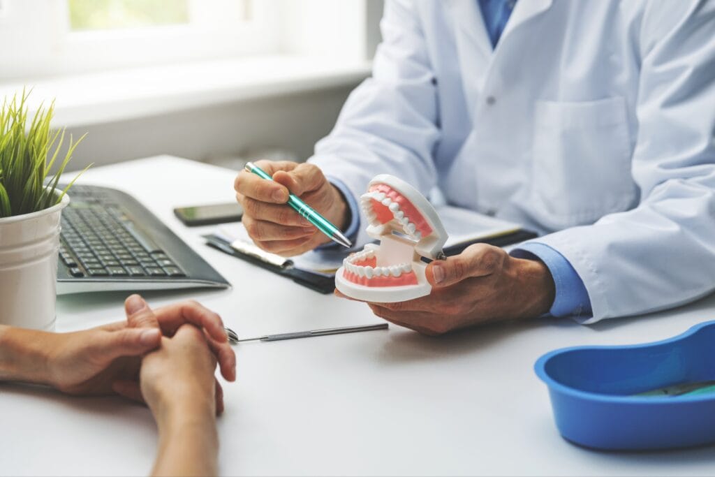 Dentist in a white coat holding a dental model and using a pen to explain a treatment plan to a patient during a full mouth restoration consultation at a desk.