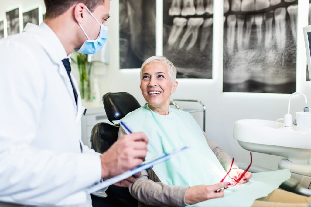 Smiling older woman sitting in a dental chair holding eyeglasses, engaging in a consultation with a male dentist wearing a face mask, with dental X-rays displayed on the wall behind them