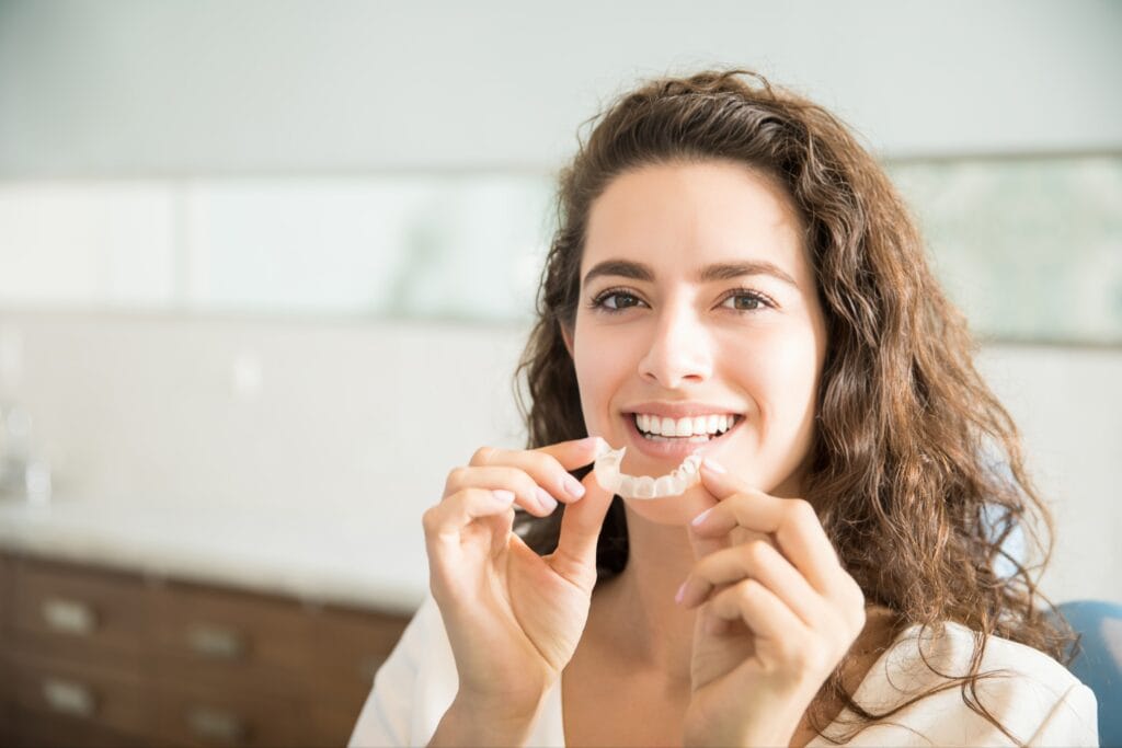 Smiling woman holding a clear Invisalign aligner near her mouth, seated in a dental office with natural lighting in the background.