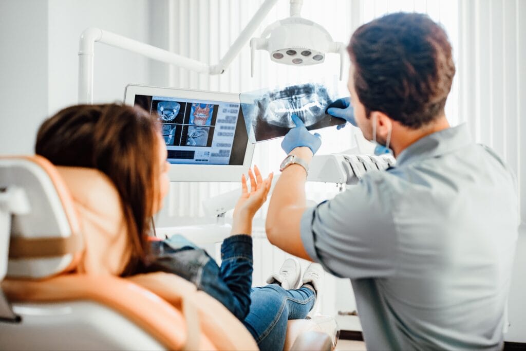 Dentist showing a dental X-ray to a female patient reclining in a chair, with additional diagnostic images displayed on a nearby monitor during a jaw surgery consultation.