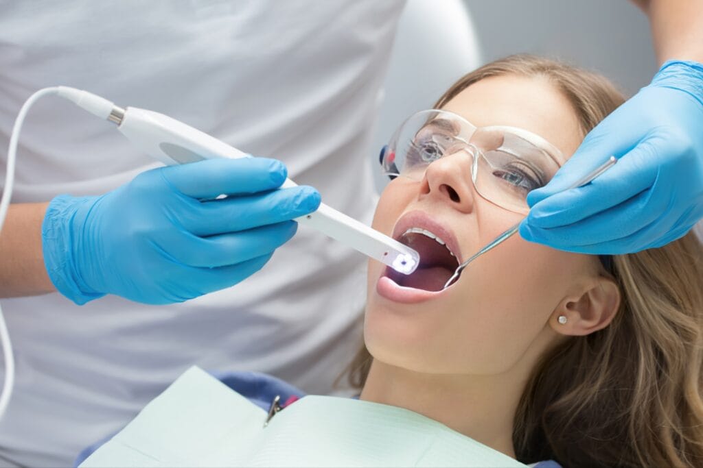 Female patient wearing protective glasses undergoing a dental examination, with a dentist using an intraoral camera and dental mirror to inspect her mouth.