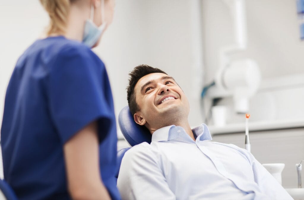 Smiling male patient reclining in a dental chair, looking up at a dental professional in blue scrubs and a face mask during an oral surgery consultation.