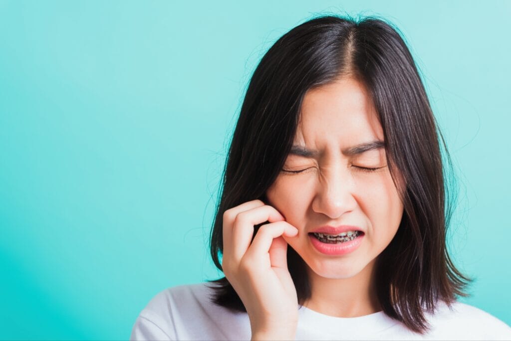 Young woman with braces wincing in pain and touching her cheek, indicating discomfort or injury, against a light blue background.