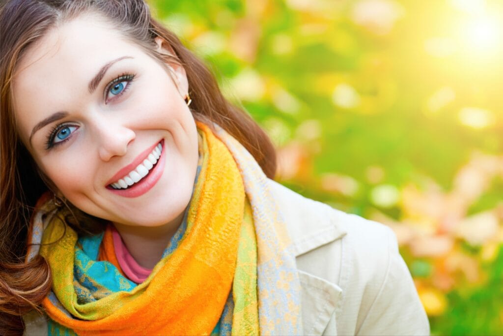 Close-up of a smiling woman with bright white teeth and blue eyes, wearing a colorful scarf and light jacket outdoors with a blurred autumn background and sunlight.
