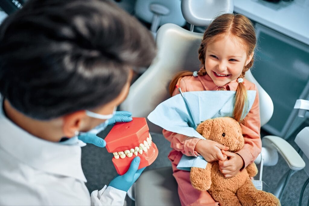 Smiling young girl with pigtails sitting in a dental chair holding a teddy bear, while a dentist shows her a model of teeth during a pediatric dental visit.