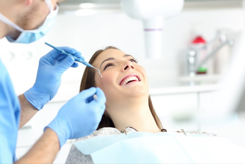 Smiling female patient reclining in a dental chair while a dentist in gloves and a mask examines her teeth with dental instruments during a routine checkup.
