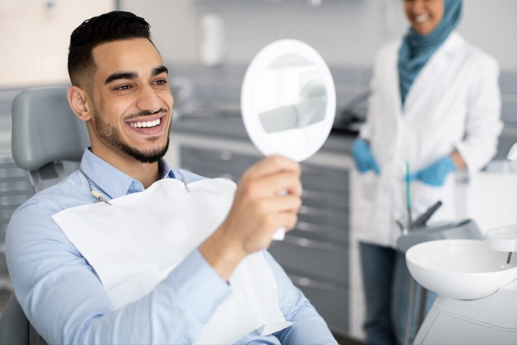 Young male patient smiling while looking at his reflection in a handheld mirror at the dental office, with a female dentist in a hijab smiling in the background.