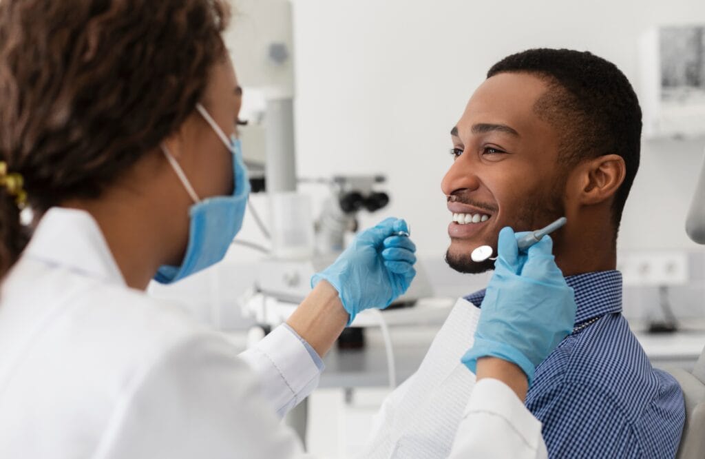 Male patient smiling in a dental chair during a reconstructive dental consultation with a masked and gloved dentist holding dental instruments.