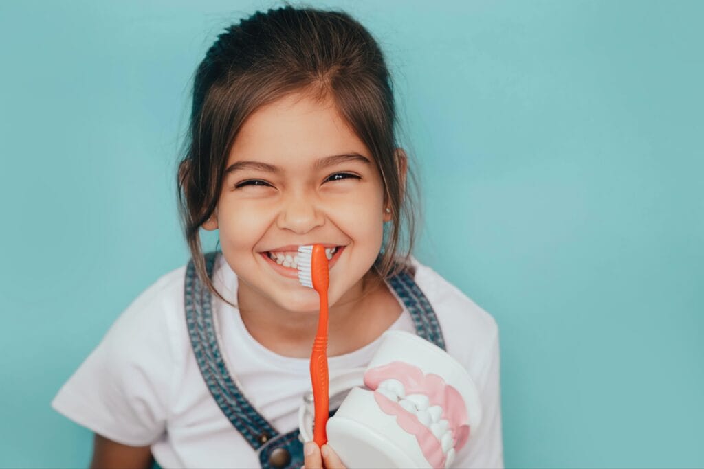 Smiling young girl holding a toothbrush and dental model, learning proper brushing technique—representing dental sealants and preventive care for children.