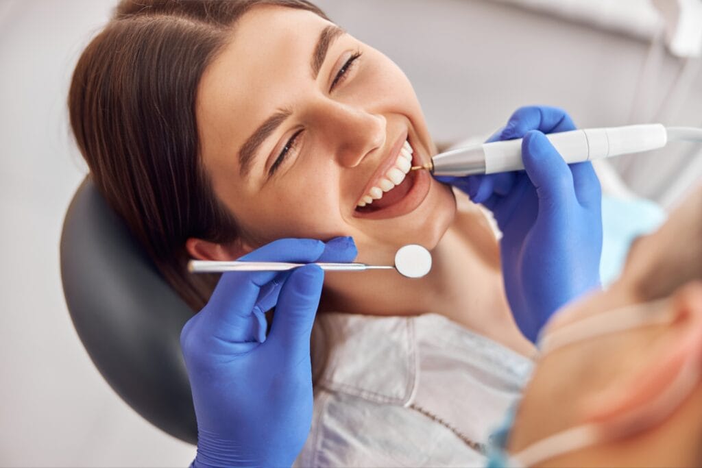 Female patient smiling while receiving a tooth contouring procedure at the dentist using a polishing tool and mirror.
