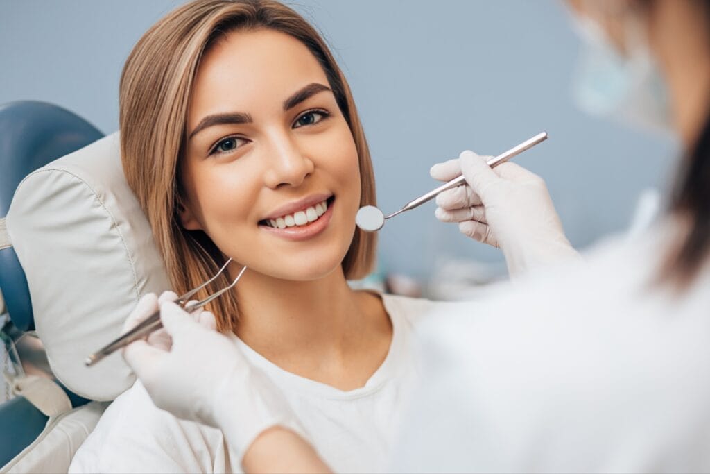 Young woman smiling in a dental chair while a dentist prepares to examine her teeth with dental tools.